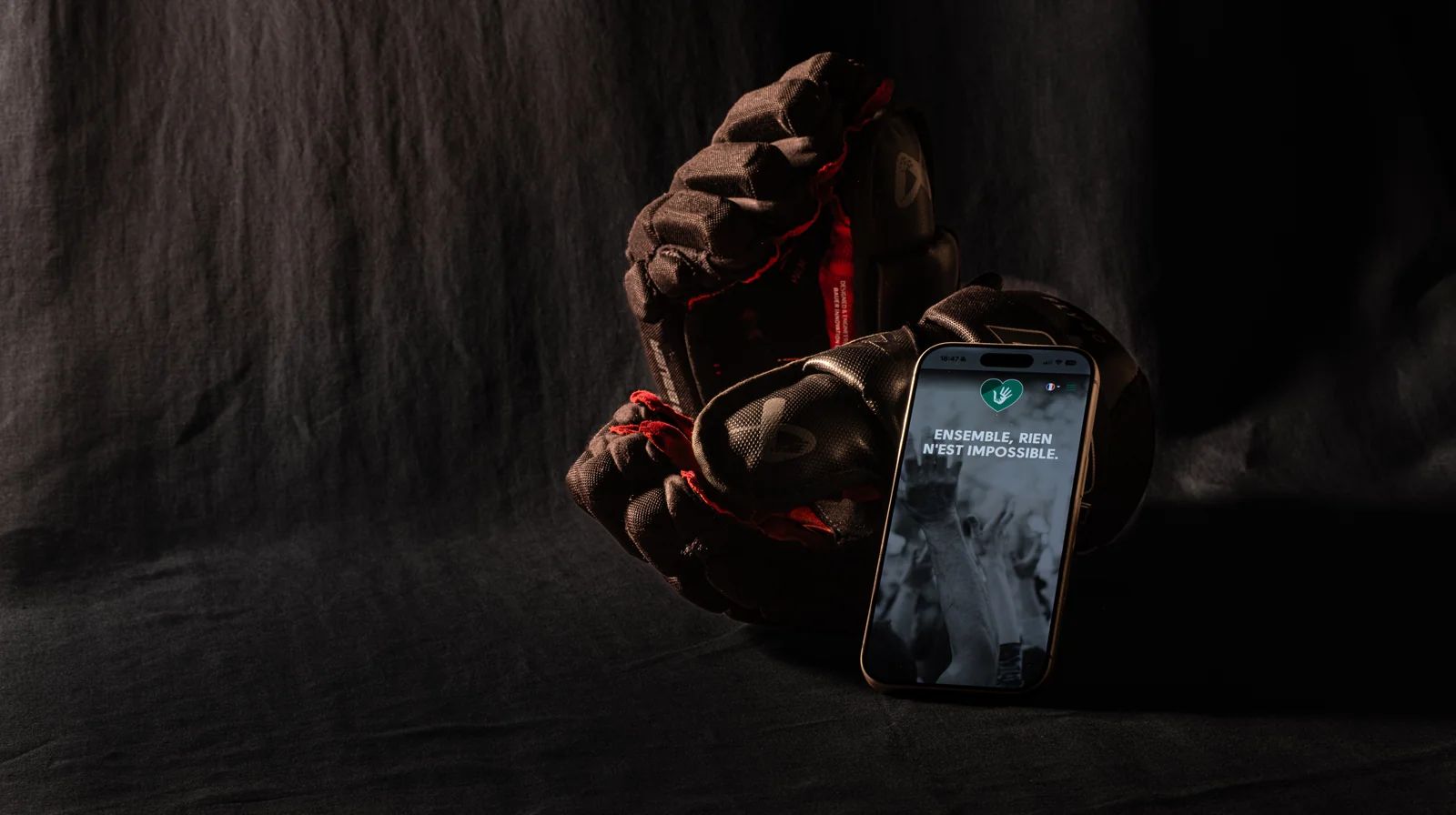 Plateforme web d’enchères caritatives UnitedHeroes, sport et caritatif : des gants de hockey éclairés d’une touche rouge, un iPhone affiche la plateforme et son mot d’ordre « Ensemble, rien n’est impossible »